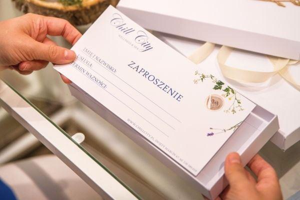 Dłonie trzymające eleganckie zaproszenie upominkowe z delikatną typografią i kwiatowymi wzorami.